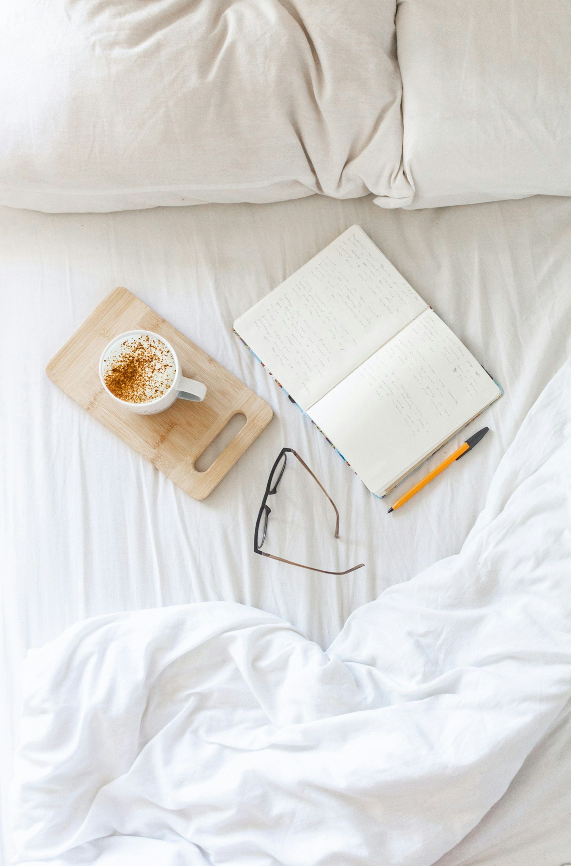 Cup of coffee on a wooden tray with a notebook and glasses on a bed.